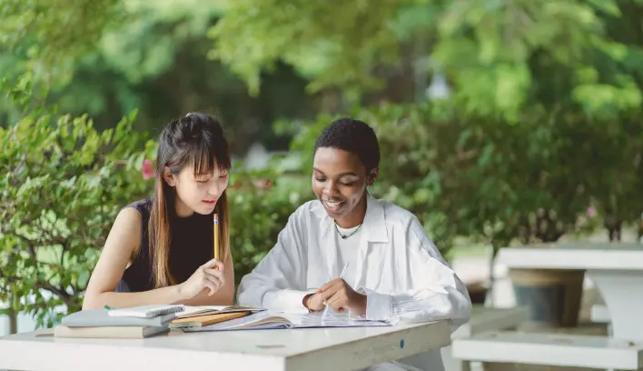 Two women studying together outside and smiling. They are surrounded by greenery.